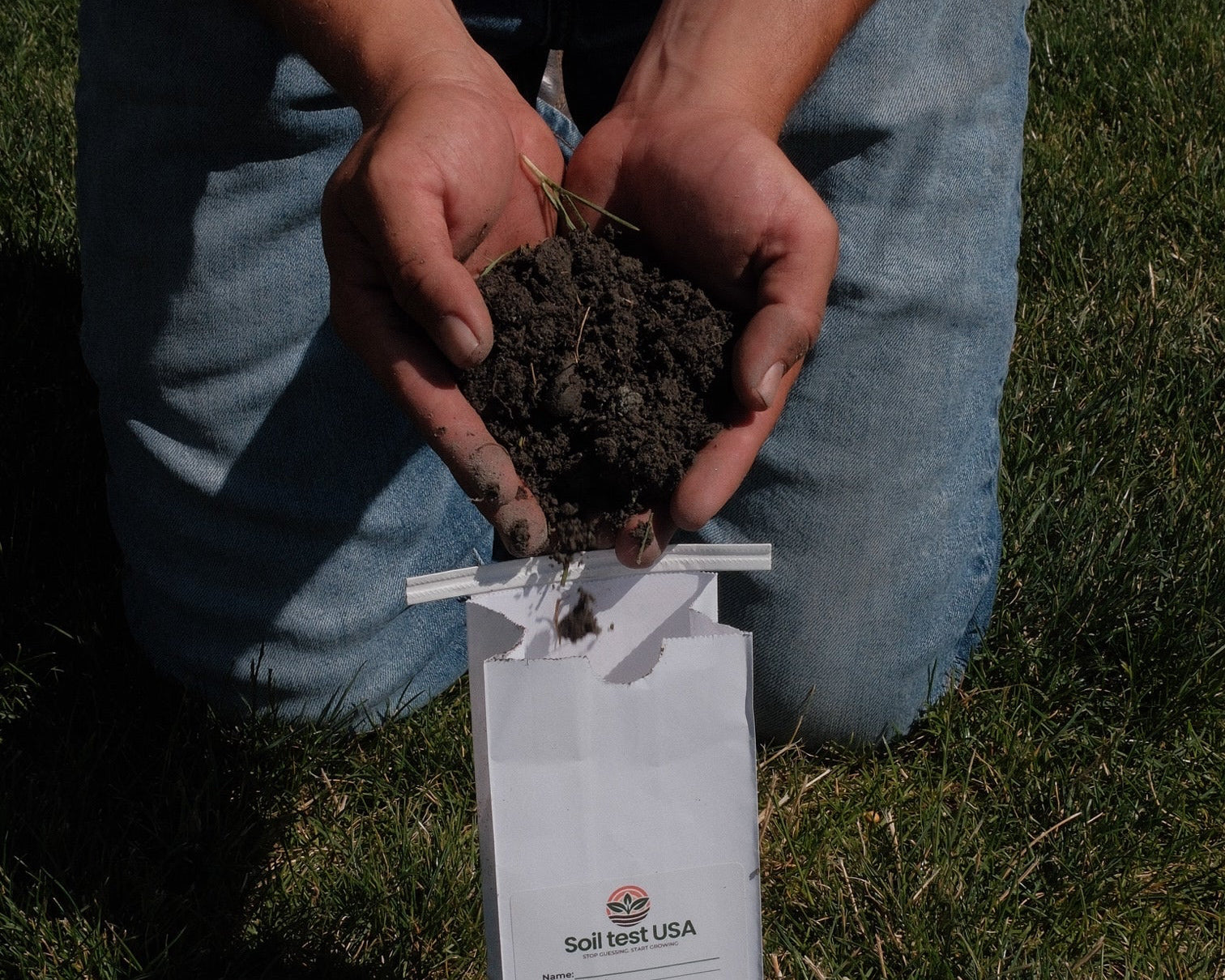 A person kneeling on grass holds a handful of dark soil next to a soil sample bag outdoor